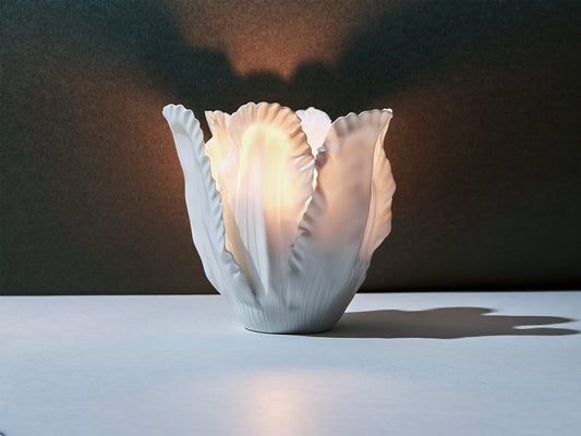 Biskuitporzellan-Windlicht aus überlappenden, blattartigen Segmenten; von innen warm erleuchtet, mit weichem Lichtschein und Schattenwurf.
Bisque-porcelain tea-light with overlapping, leaf-like segments; softly lit from within, casting a warm glow and delicate shadows.