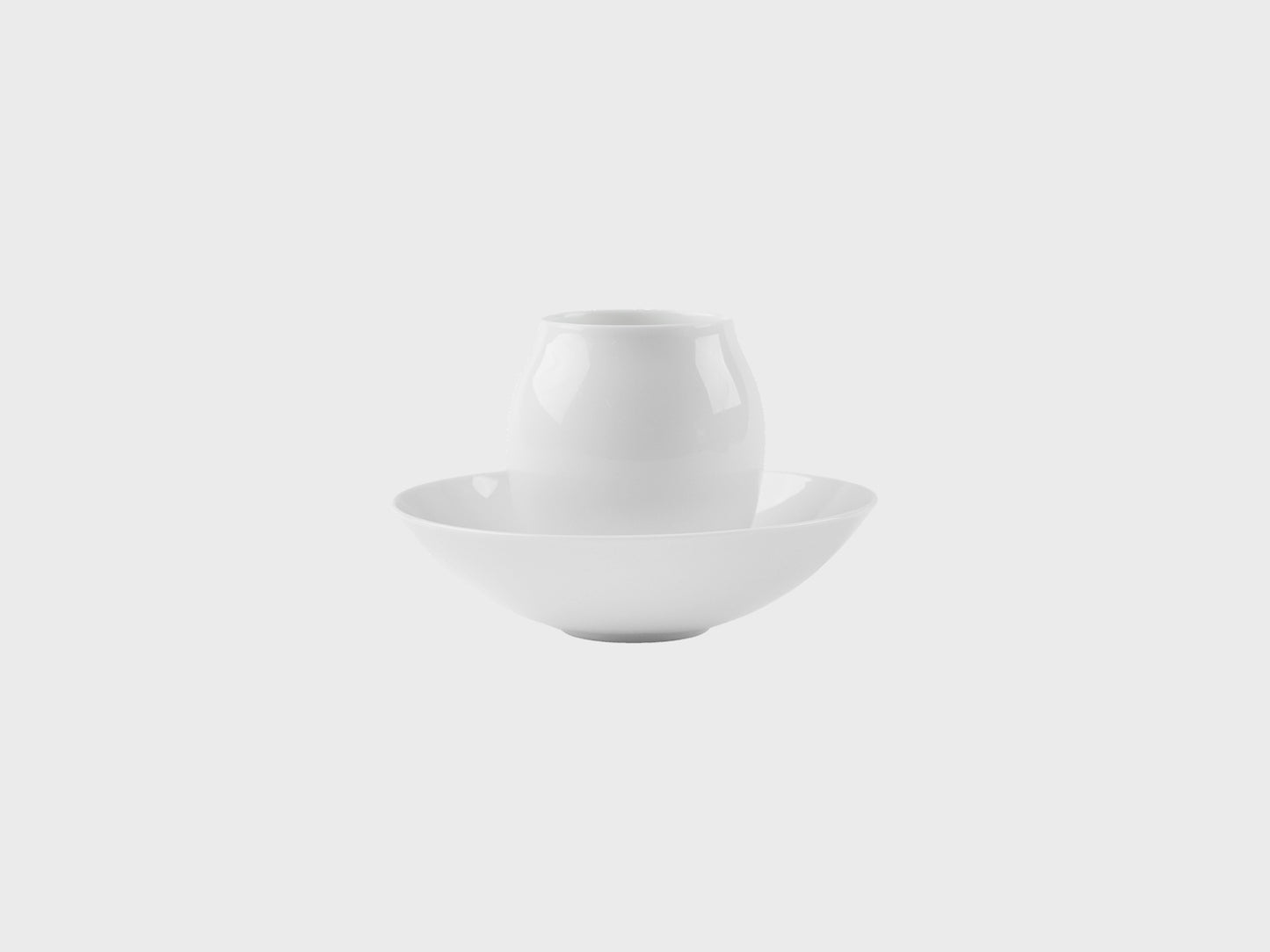 Zwei weiße Porzellanobjekte auf hellem Hintergrund: eine kleiner, bauchiger Mokka Becher steht mittig in einer flachen Schale. Beide Objekte sind schlicht, glänzend glasiert und ohne Dekor.
Two white porcelain objects on a light background: a small, rounded mocca beaker stands centered inside a shallow bowl. Both pieces are simple, glossy glazed, and undecorated.