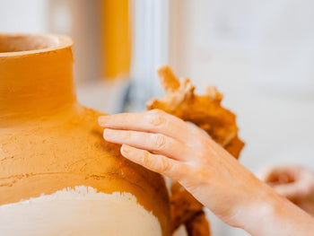 Detailaufnahme der Hände der Künstlerin Nigin Beck, die ein großes Gefäß aus Ton modellieren. Die Oberfläche zeigt raue, organische Strukturen und teilweise weiß grundierte Bereiche.

Close-up of artist Nigin Beck's hands shaping a large clay vessel. The surface features rough, organic textures and partially white-primed areas.
