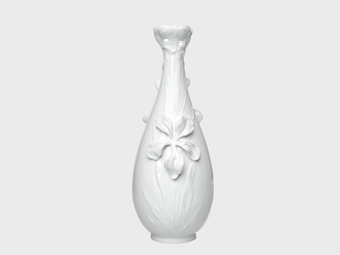 Weiße Porzellanvase mit glänzender Oberfläche und floralen Reliefdetails auf neutral hellgrauem Hintergrund. Die bauchige Form wird durch eine große Blütenapplikation auf der Vorderseite und eine dekorativ geformte, blütenartige Öffnung am Hals ergänzt.

White porcelain vase with a glossy finish and floral relief details on a neutral light gray background. The rounded body features a large flower applique on the front, while the neck is finished with a decorative, blossom-shaped opening.
