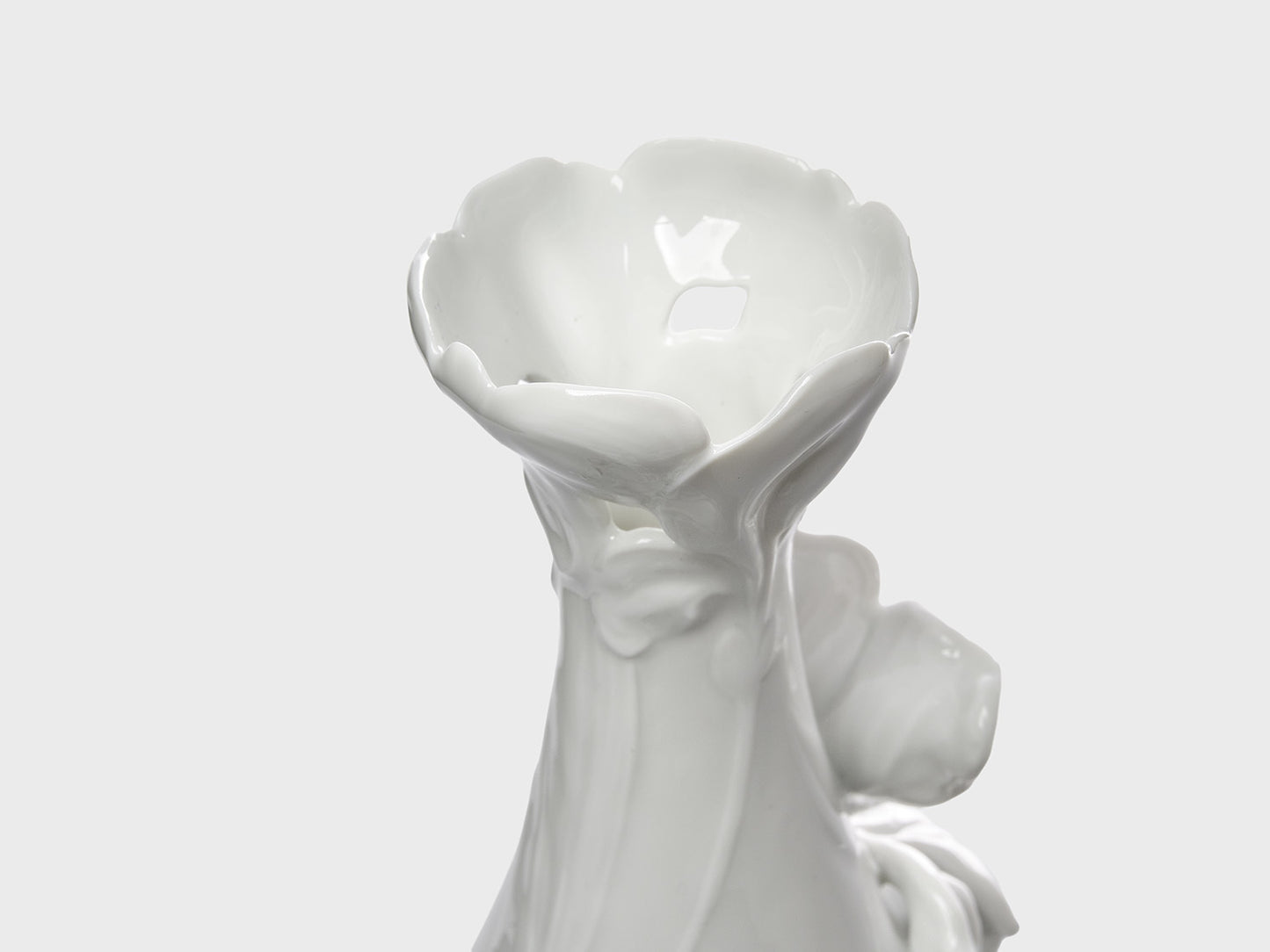 Nahaufnahme der blütenförmig gestalteten Öffnung einer weißen Keramikvase mit glänzender Oberfläche. Die organisch geformten Blütenblätter und feinen Reliefdetails unterstreichen das florale Design vor einem neutral hellgrauen Hintergrund.

Close-up of the blossom-shaped opening of a white ceramic vase with a glossy finish. The organically sculpted petals and subtle relief details highlight the floral design against a neutral light gray background.