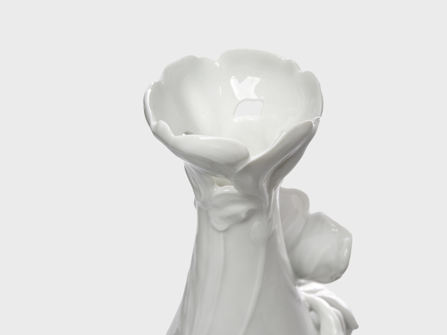Nahaufnahme der blütenförmig gestalteten Öffnung einer weißen Keramikvase mit glänzender Oberfläche. Die organisch geformten Blütenblätter und feinen Reliefdetails unterstreichen das florale Design vor einem neutral hellgrauen Hintergrund.

Close-up of the blossom-shaped opening of a white ceramic vase with a glossy finish. The organically sculpted petals and subtle relief details highlight the floral design against a neutral light gray background.