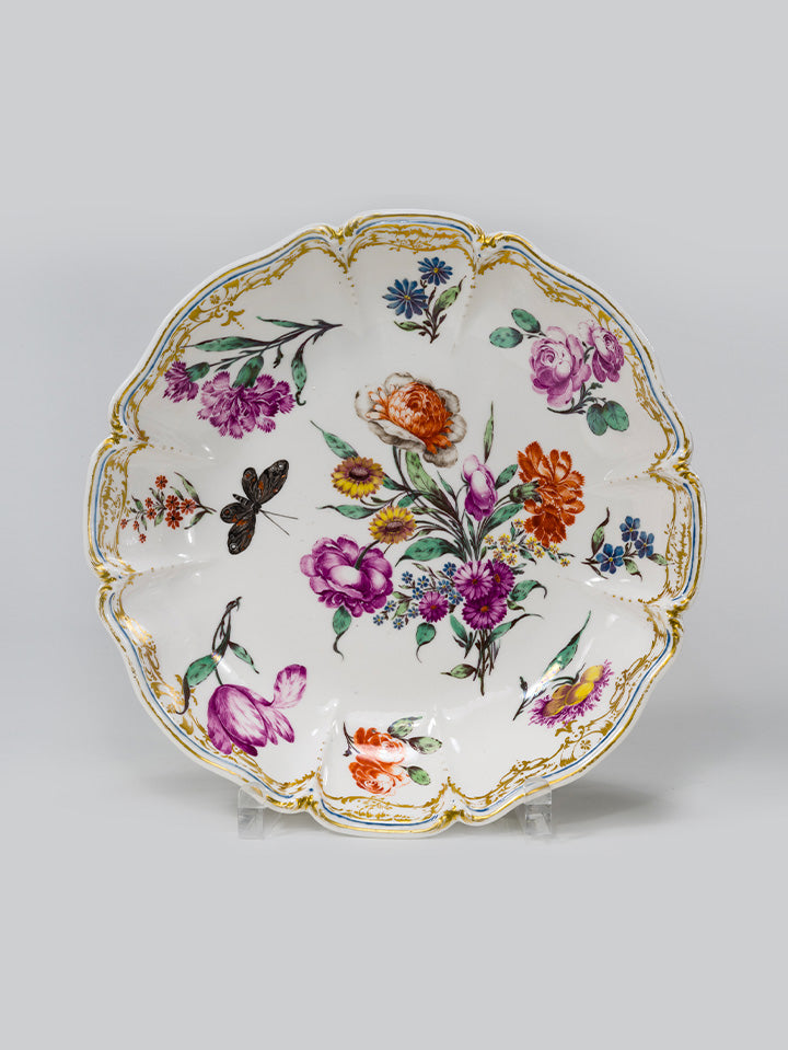 Barocker Teller mit floralem Dekor, goldverziertem Rand und schwarzem Schmetterling.

Baroque plate with floral décor, gilded rim, and black butterfly.
