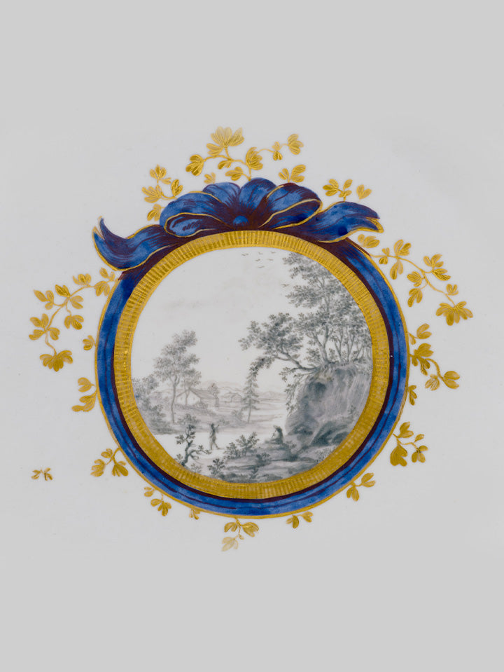 Detailansicht eines Porzellanmedaillons mit feiner Landschaftsmalerei in Schwarz-Weiß, umrahmt von kobaltblauer Schleife und Golddekor.

Close-up of a porcelain medallion with fine black-and-white landscape painting, framed by cobalt-blue ribbon and gold decoration.