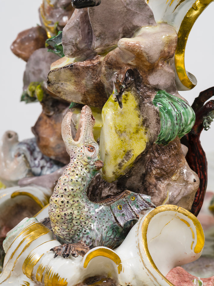 Phantasievolles, echsenartiges Tier mit aufgerissenem Maul und detailreich bemaltem Körper, modelliert zwischen Felsen und Porzellanschnecke.

Fantastic lizard-like creature with gaping mouth and intricately painted body, positioned among rocks and a porcelain snail.