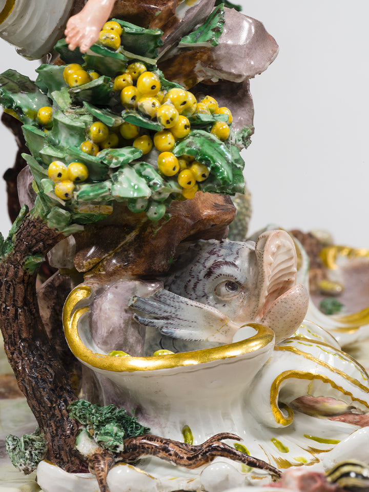 Gelbe Porzellanbeeren auf grünem Blattwerk neben einem fein modellierten Fischkopf, teils hinter Blattwerk versteckt.

Yellow porcelain berries on leafy greenery beside a delicately modelled fish head, partially hidden by foliage.